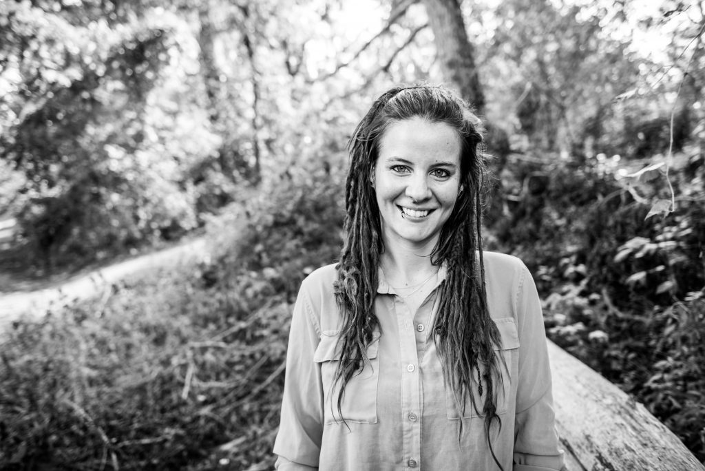 Black & white photo of Rachel, a white woman with dark dreadlocks wearing a button-down top, standing outdoors.
